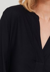 Haut en tissu noir avec un décolleté en V, présentant une texture lisse, des détails de coutures subtils et une coupe décontractée au niveau des épaules.