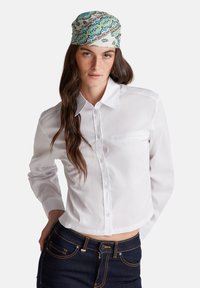 Camicia bianca con bottoni e maniche lunghe, dotata di tasca. Indossata con jeans in denim scuro. Il foulard per la testa presenta un motivo floreale colorato su sfondo chiaro.