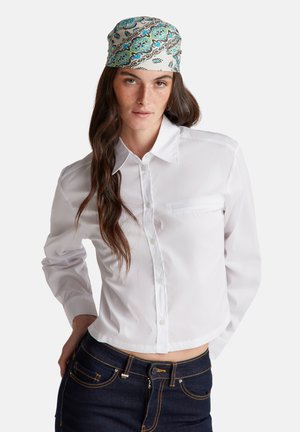 Chemise blanche à boutons, à manches longues, avec une poche. Portée avec un jean en denim foncé. Le foulard est à motifs floraux colorés sur fond clair.