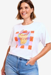 Šypsosi moteris, vilkinti baltą marškinėlį su ryškiu grafiku, vaizduojančiu aštrų margaritos gėrimą, ir tekstu, derinama su mėlynomis džinsinėmis kelnėmis, ranka kišenėje.