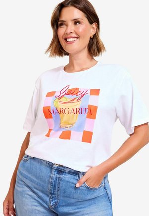 Lächelnde Frau, die ein weißes T-Shirt mit buntem Grafikdesign eines scharfen Margarita-Getränks und Text trägt, kombiniert mit blauen Jeans, die Hand in der Tasche.