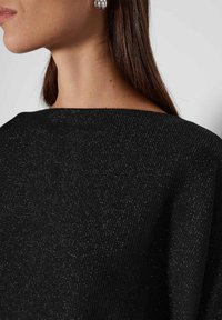 Schwarzes Strickoberteil mit weitem Ausschnitt, das einen subtilen metallischen Schimmer aufweist. Die Textur wirkt gerippt, und der Stoff fällt weich.