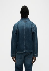 Μπουφάν τζιν σε σκούρο μπλε, με κλασικό γιακά, μακριά μανίκια και ορατές ραφές, σετ με τζιν παντελόνια στο ίδιο χρώμα.