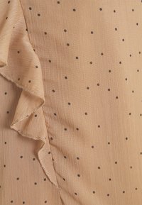 Tissu beige avec de petits pois marron foncé, présentant un bord froncé sur le côté gauche. La texture semble lisse avec un léger éclat.