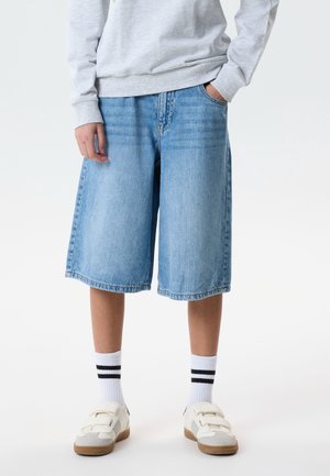 Person iført lyseblå denim shorts til knæet, hvide sneakers med velcrolukning, hvide sokker med sorte striber og en lysegrå sweatshirt.