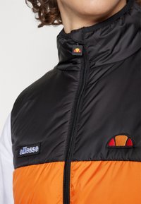 Černý a oranžový péřový vest se zipem, vysokým límcem a vyšitým logem. Hladká textura s barevným blokovým designem.