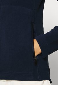 Giacca in pile blu navy con tasca laterale con zip, texture liscia e silhouette aderente, colletto alto e maniche lunghe.