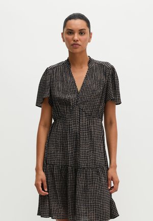 Donna con capelli scuri raccolti indossa un vestito nero a maniche corte con pois beige, in piedi davanti a uno sfondo chiaro uniforme.
