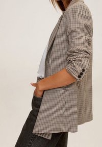 Femme portant un blazer pied-de-poule beige et marron avec les manches retroussées et des boutons noirs sur un haut blanc, une main dans la poche de son jean foncé.