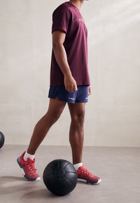 Zapatillas deportivas rojas y navy, camiseta marrón, pantalones cortos navy. De pie junto a una pelota de ejercicio negra sobre un suelo gris claro.