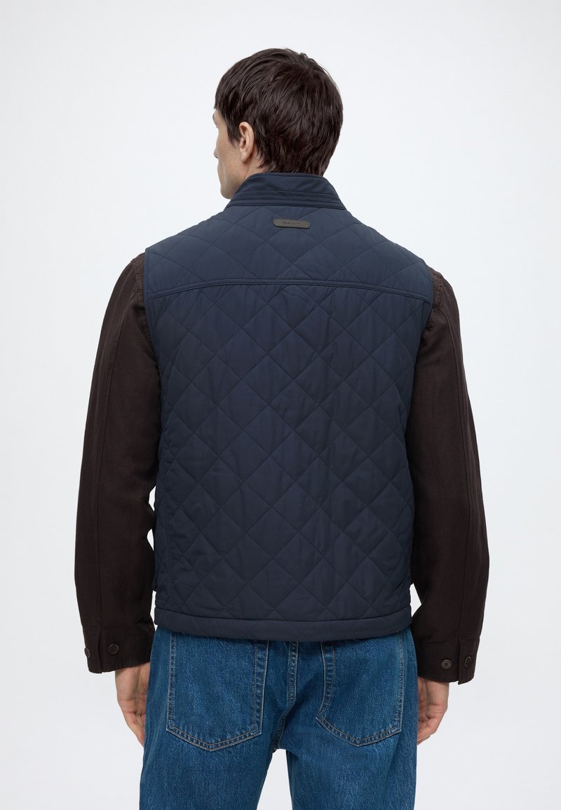 Mand iført en marineblå vatteret vest over en brun langærmet skjorte og blå jeans, vendt væk mod en ensfarvet lys baggrund.