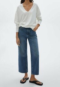 Top bianco a maniche lunghe con scollatura a V abbinato a jeans blu a gamba larga a vita alta. Il modello indossa ciabatte nere e mostra una vestibilità rilassata.