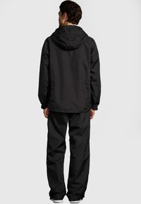 Svart vattentät jacka med huva, med avslappnad passform, elastiska ärmslut och matchande byxor. Strukturerad tyg med minimal detaljering.