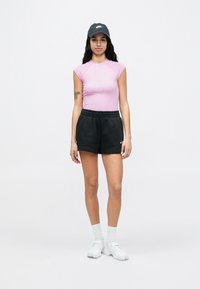 Nő, aki fekete Nike sapkát, rózsaszín ujjatlan felsőt, fekete Nike sortot, fehér zoknit és fehér cipőt visel, és egyszínű háttér előtt áll.