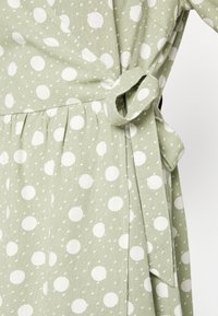 Grön omslagklänning i texturerat tyg med stora vita prickar. Har ett bälte vid midjan och plisserad kjoldetalj.
