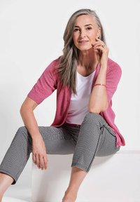 Rosa gestrickte Strickjacke mit kurzen Ärmeln, über einem weißen Oberteil getragen. Schwarz-weiß karierte Hose in schlanker Passform. Neutraler Hintergrund.