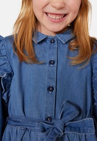 Jeanskleid in Hellblau mit Kragen, Knopfleiste, Rüschsäumen und gebundenem Taillendetail. Glatte Textur und klassisches Design.