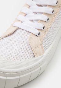 Sneaker branco e bege com parte superior em malha texturizada, atacadores de algodão e sola de borracha com estrias horizontais distintas. Ilhoses prateados acentuam o design.