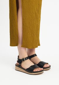 Czarne sandały na koturnie z odkrytymi palcami, skórzanymi paskami i złotą klamrą. Podeszwa ma drewnianą teksturę oraz czarne przeszycia.