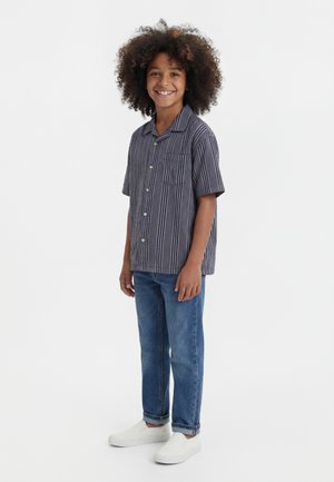 Ένα παιδί με κατσαρά μαλλιά και χαμόγελο φοράει μια κοντομάνικη ριγέ πουκάμισο με κουμπιά, μπλε τζιν και λευκά παπούτσια χωρίς κορδόνια, ενώ στέκεται μπροστά σε λευκό φόντο.