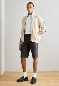 Beige ribbad zip-tröja över en vit t-shirt, ihop med grå denimshorts och svarta slip-on skor med tjock sula.