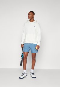Hvit hettegenser, lyseblå shorts, holder en tenniskølle og ball. Hvite joggesko med marineblå detaljer, kort frisyre.