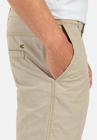 Beige chinos met schuine zakken en een achterzak. Kenmerkt zich door een subtiel bruin logo nabij de achterzak en zichtbare stikseldetails.