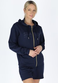 Navy zip-up hoodie with a gold zipper, hood, and front print reading “SEASIDE.” Soft material and ribbed cuffs and hem. Paired with shorts.