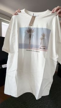 Weißes T-Shirt mit Aufdruck von Palmen, einem weißen Van und dem Text "VON SOMMER TRÄUMEN", gehalten von Händen drinnen mit einem angehängten Kleidungsetikett.