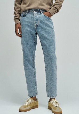 Personne portant un jean droit bleu clair, un pull beige et des baskets beiges à lacets blancs, debout avec une main dans la poche.