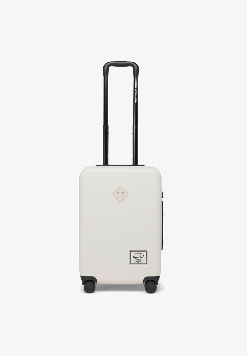 Valise rigide de couleur crème avec une surface texturée, poignée télescopique noire, quatre roues et un patch logo à l'avant.