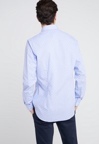 Camisa azul claro con pequeños lunares rojos, de manga larga, con puños abotonados y dobladillo curvado. Tejido suave con cuello tipo polo.