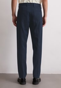 Pantalon bleu marine en mélange de coton, à coupe droite, avec deux poches arrière et des coutures nettes et ajustées. Chaussures noires visibles en bas.
