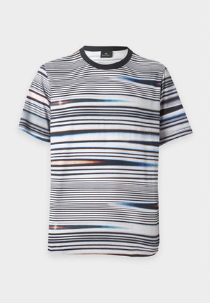Mann steht mit Händen in den Taschen, trägt gestreiftes Kurzarmhemd, weiße Hose und gelbe Slip-On-Schuhe vor weißem Hintergrund.
