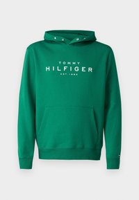 Zelená mikina s kapucí vyrobená z bavlny, která má přední klokankovou kapsu, stahovací kapuci a bílý text s logem "TOMMY HILFIGER EST. 1985" na hrudi.
