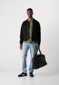 Black bomber jacket over a green shirt, light blue jeans, and black sneakers. Carrying a large black leather bag. Plain background.