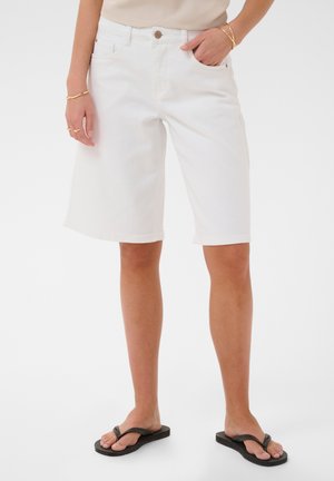 Kvinde iført hvide knælange jeansshorts, beige top, sorte klipklappere og gyldne armbånd og ringe, står med den ene hånd i lommen.