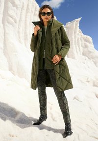 Olijfgroen, gewatteerde lange pufferjack; hoge kraag, gedragen over een bijpassende top en gemusterde, donkergroene slangenprintsokken; zwarte enkellaarsjes.