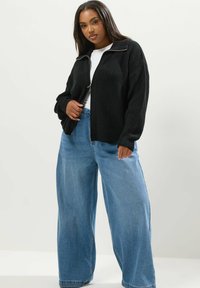 Svart stickad zip-jacka med ribbat mönster, ihop med ljusblå vida jeans. Enkel vit t-shirt under.