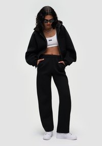 Sort zip-up hættetrøje lavet af blødt stof, parret med en hvid cropped tank top og sorte sweatpants med en afslappet pasform og lommer.