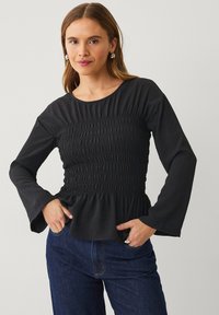 Zwarte langetermijn top met smockontwerp bij de taille en uitlopende mouwen. Zachte textuur, ronde halslijn en peplumzoom. Gepaarde met denim.