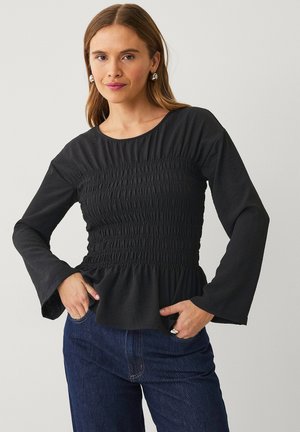 Haut noir à manches longues avec un design smocké à la taille et des manches évasées. Texture douce, col rond et ourlet peplum. Associé avec du denim.