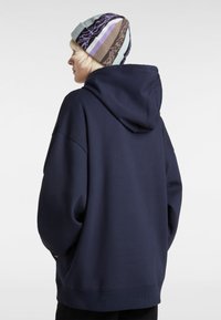 Hoodie oversize bleu marine avec une grande poche à l'arrière et des poignets côtelés. Porté avec un bonnet en tricot à motifs présentant des rayures vertes, brunes et violettes.