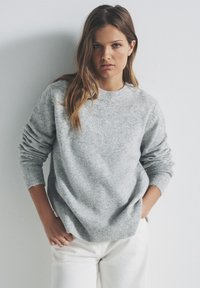 Maglione grigio chiaro in maglia con scollo rotondo, polsini e orlo a coste. Indossato con pantaloni bianchi a vestibilità rilassata su uno sfondo neutro.