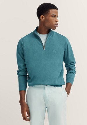 Teal zip-up sweater in soft fabric with ribbed cuffs and collar, worn over a light blue shirt, paired with light-colored trousers.