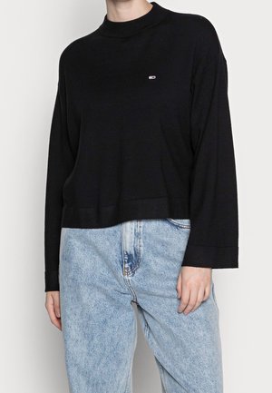 Žena oblečená v černém topu s dlouhým rukávem a malým logem na hrudi a ve světle modrých džínách s vysokým pasem, stojící před jednoduchým pozadím.