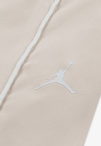 Svijetlo bež tkanina s bijelim siluetom Jordan logotipa i kontrastnim bijelim šavovima, koja prikazuje glatku teksturu i sportski dizajn.