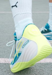 Sport shoe with a gray and white upper, green accents, textured sole, and blue patterns. Features white laces and a prominent logo.