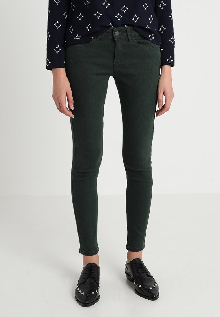 Femme portant un pantalon slim vert foncé, des chaussures noires à clous pointues, et un haut à manches longues bleu marine avec des motifs en losanges blancs.