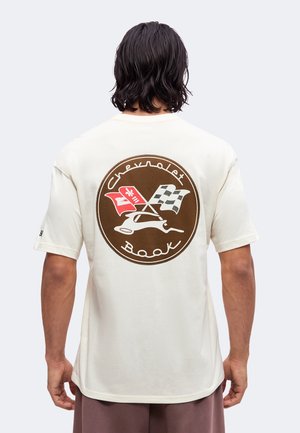 Homme aux cheveux foncés mi-longs portant un T-shirt blanc avec un logo Chevrolet marron et des drapeaux croisés dans le dos.